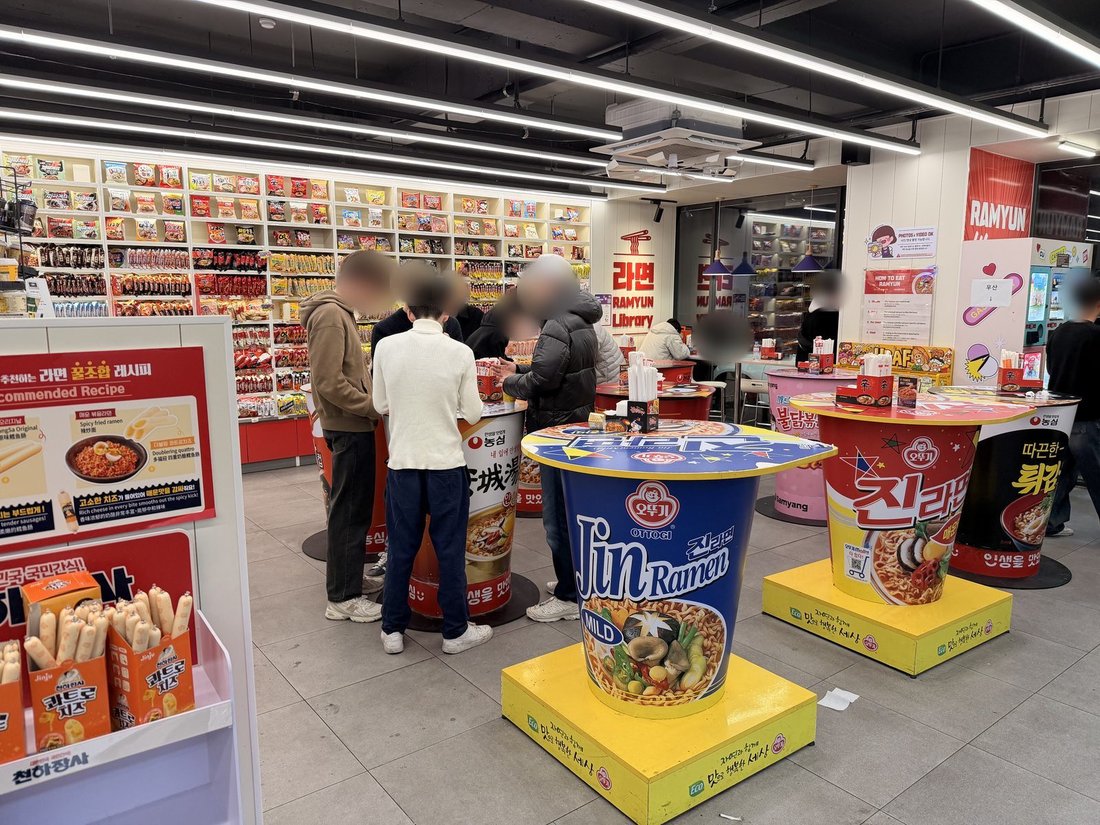 Shoppers at tables shaped like giant instant ramen cups in CU's Ramyun Library in Seoul, with a wall of ramen behind them