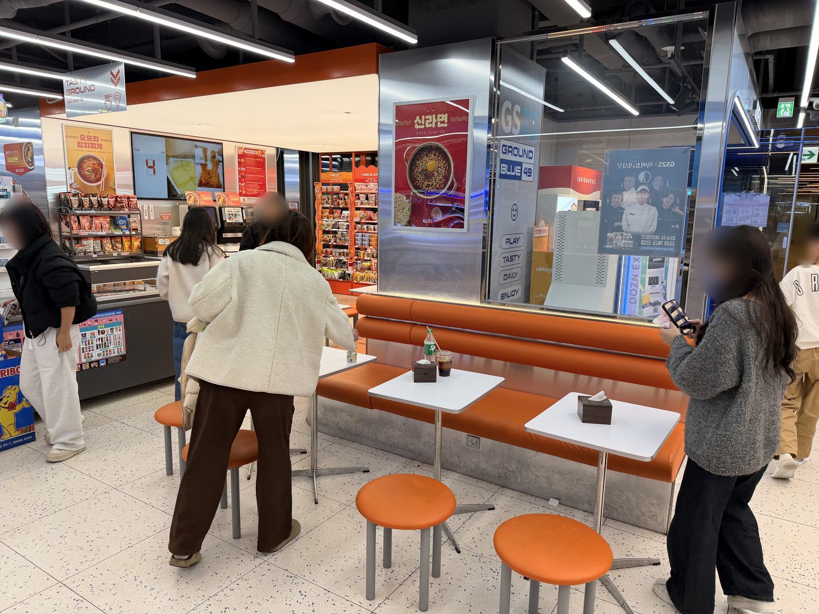 Shoppers at the tables and orange benches of a GS25 eat-in corner in Seoul, near its ramen shelves and cooking machines