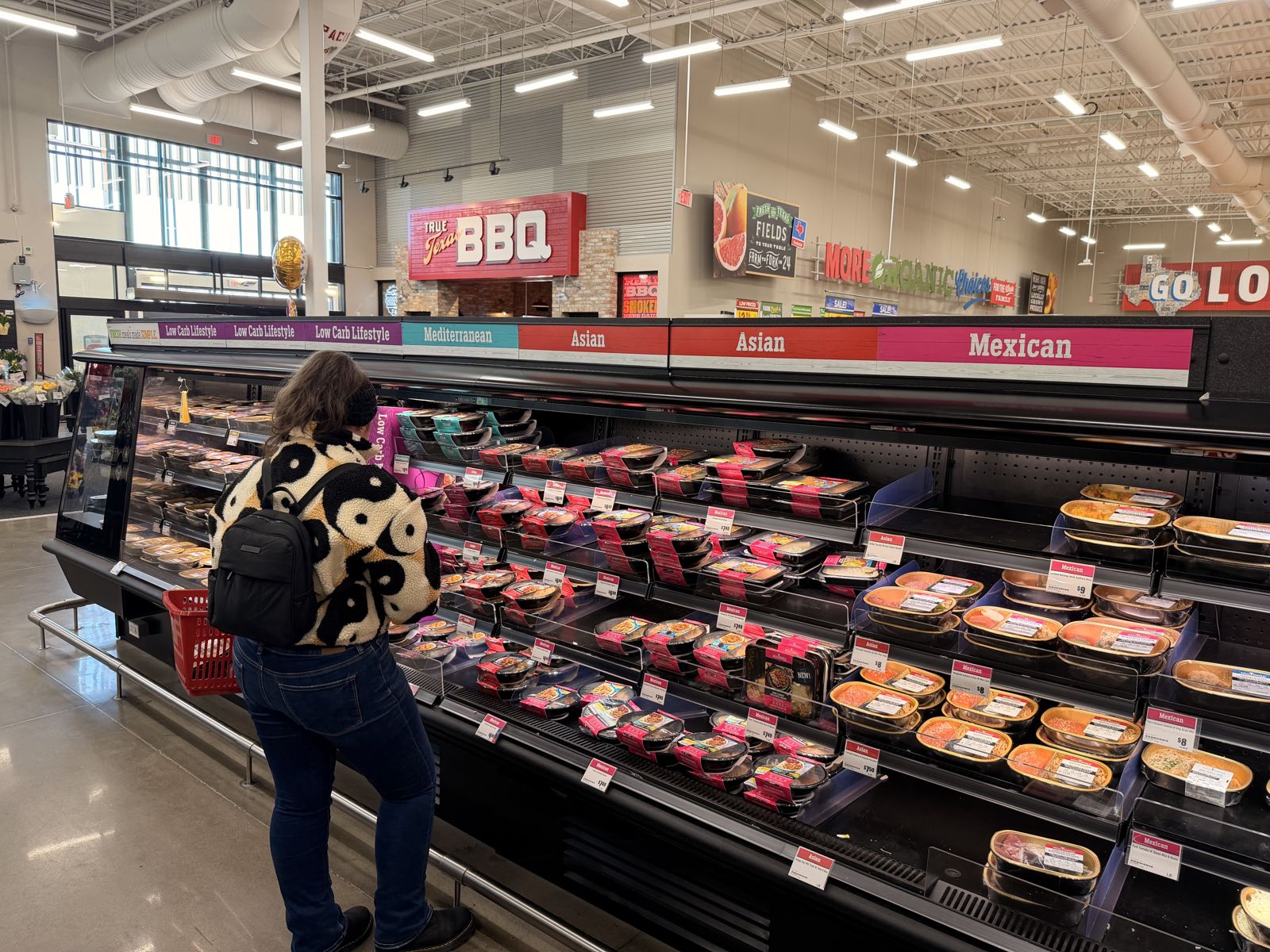 A shopper at an H-E-B prepared-meals case signed Low Carb Lifestyle, Mediterranean, Asian and Mexican