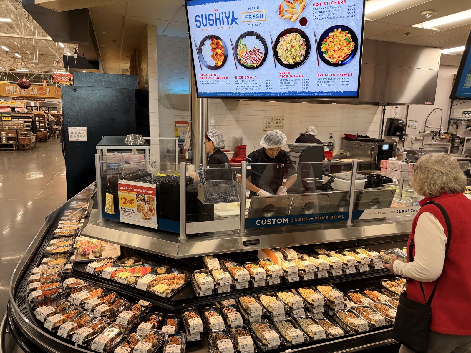 H-E-B Sushiya counter with staff making sushi under a menu board, and a shopper at the sushi case