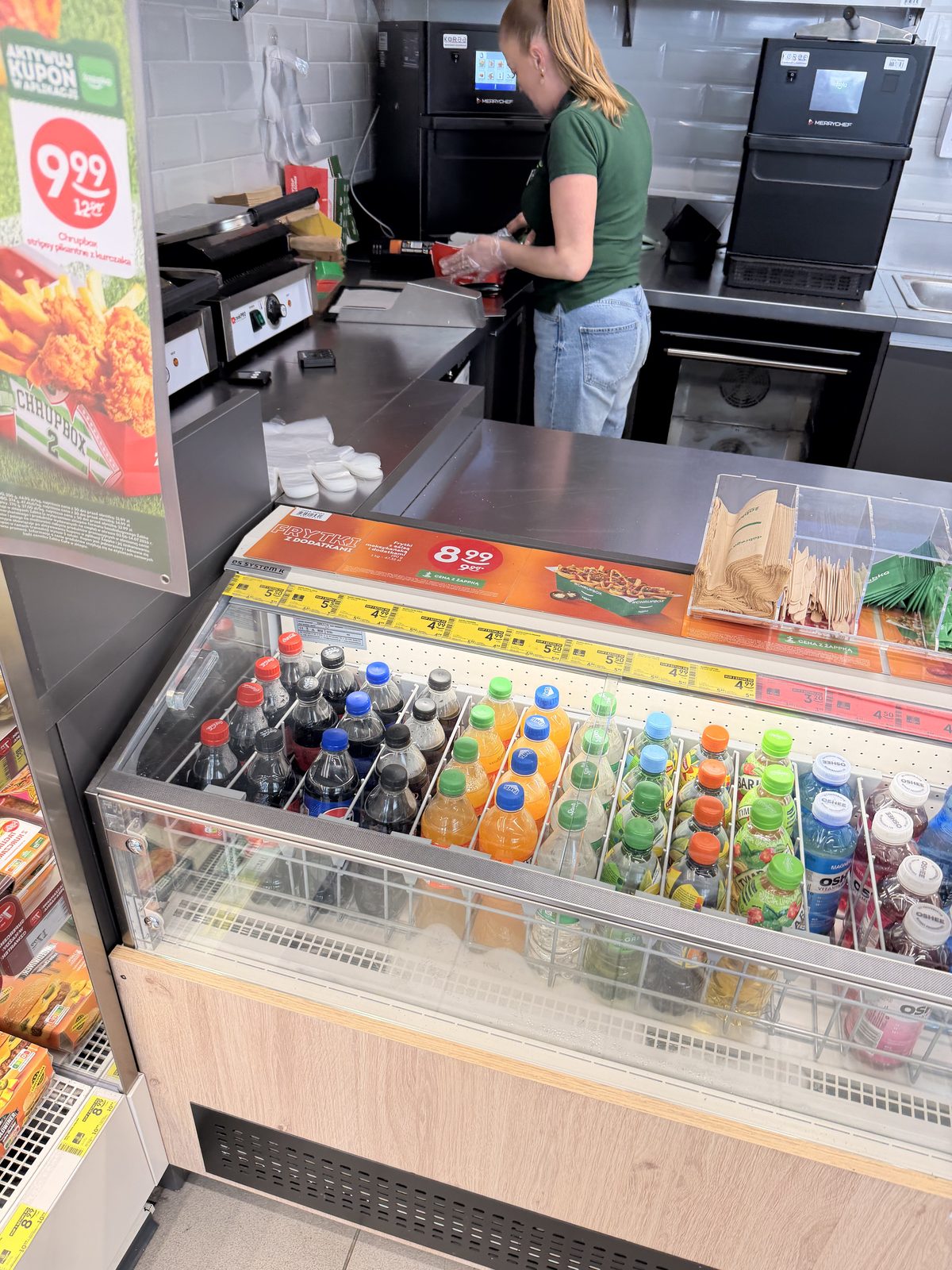 A Żabka employee at a high-speed oven behind the counter, next to a loaded fries price sign