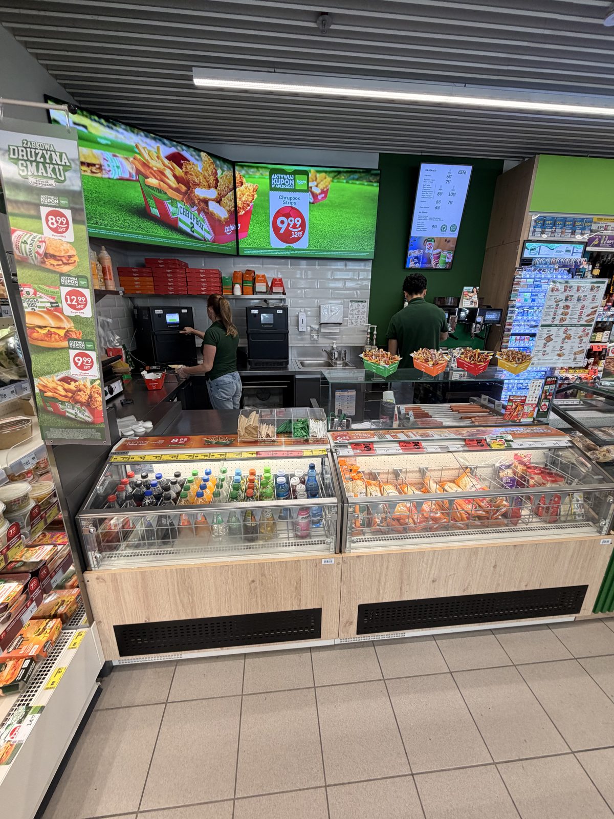 A Żabka hot food counter with staff, high-speed ovens and menu screens showing loaded fries